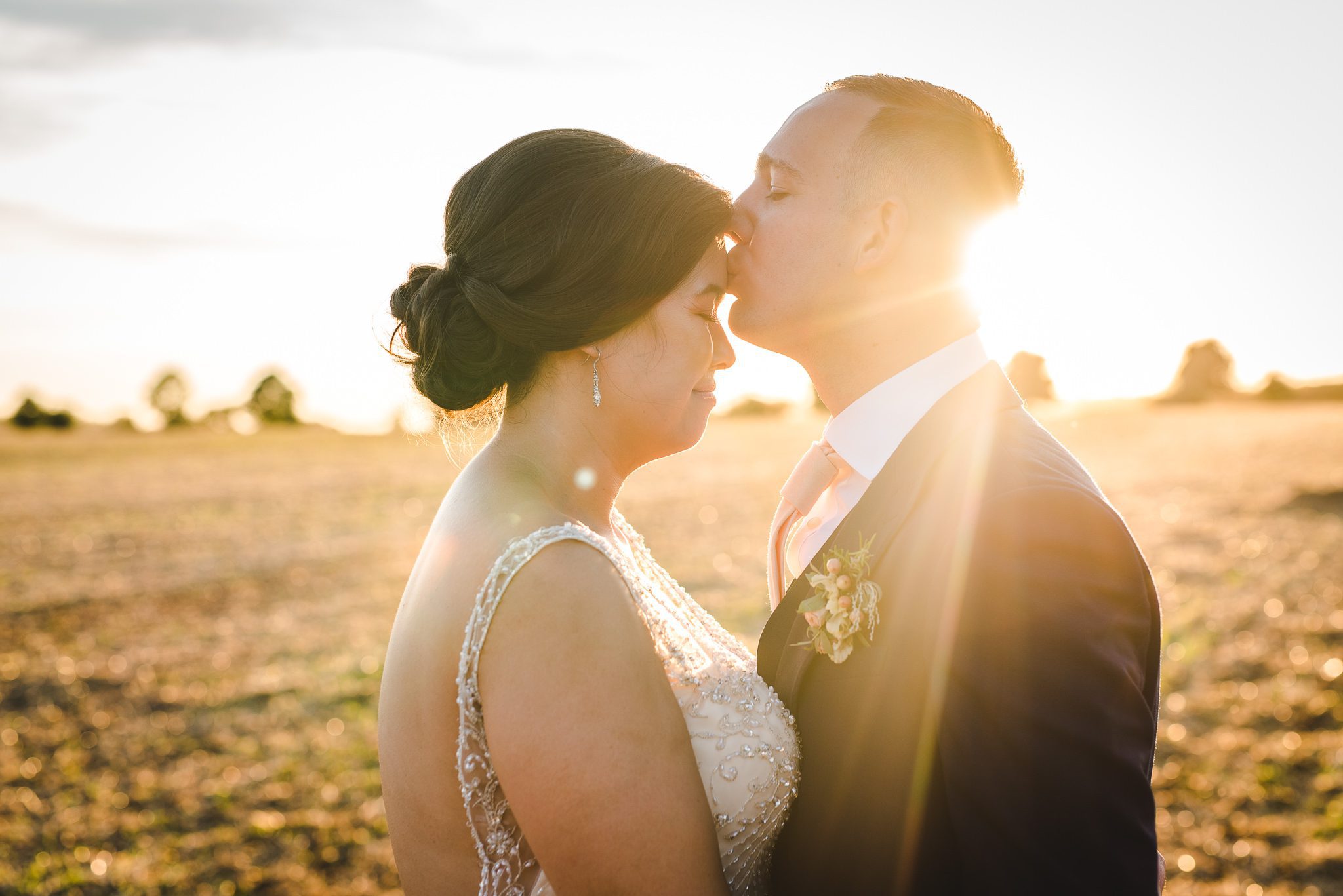 Groom kissing brides forehead in front of sunset at stone barn by Bigeye Photography
