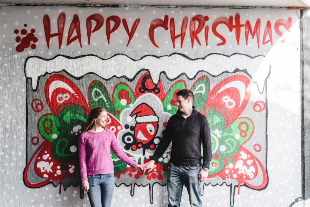A couple holding hands in front of a Happy Christmas painting